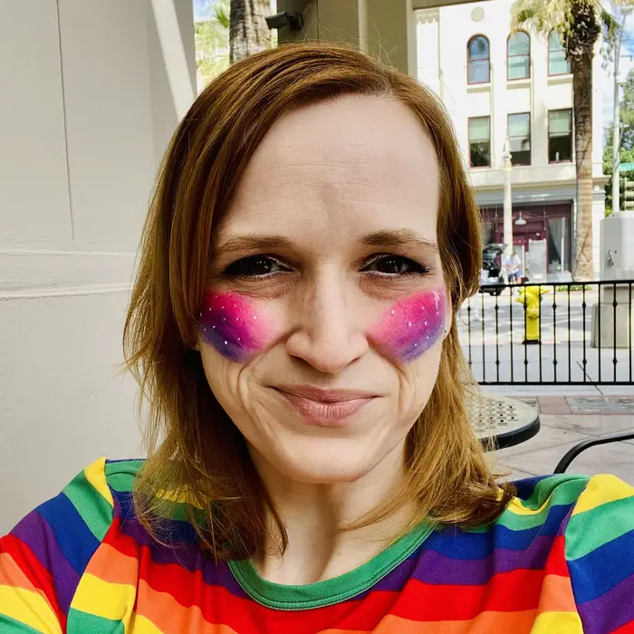 Meg in a rainbow striped shirt with pink and purple glitter on her cheeks, smiling on a city sidewalk