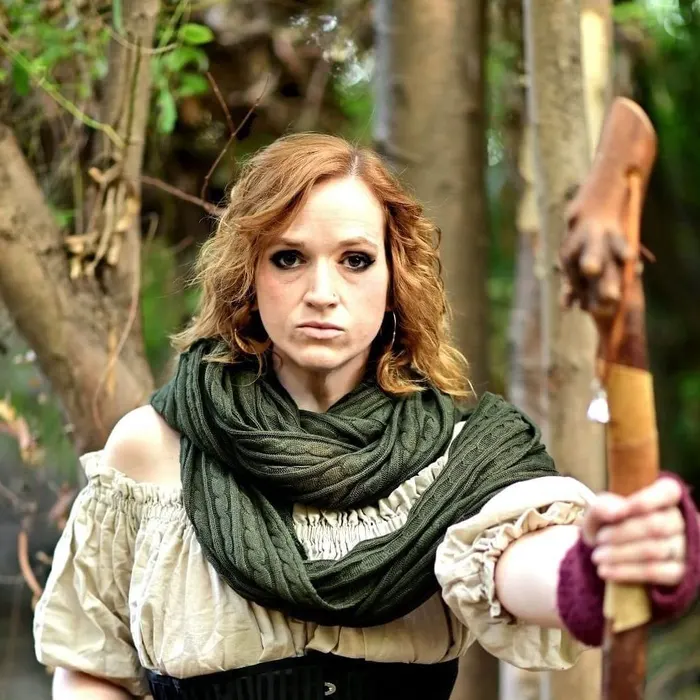 Meg in a fantasy costume with a corset, green scarf, and fingerless gloves, holding a carved wooden staff in the woods and looking very serious