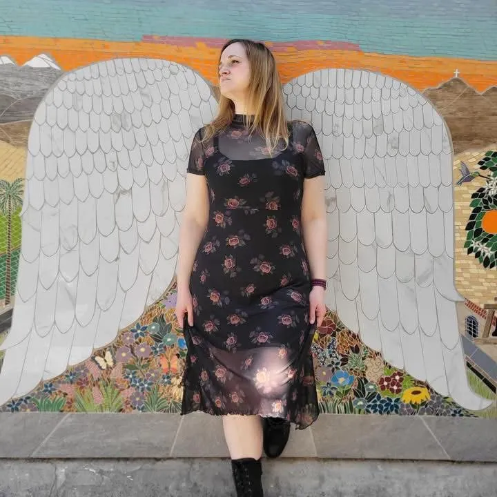 Meg in a floral mesh dress, standing in front of a mosaic mural so a pair of giant white wings appear to come from her back, chin up and gazing off to the side