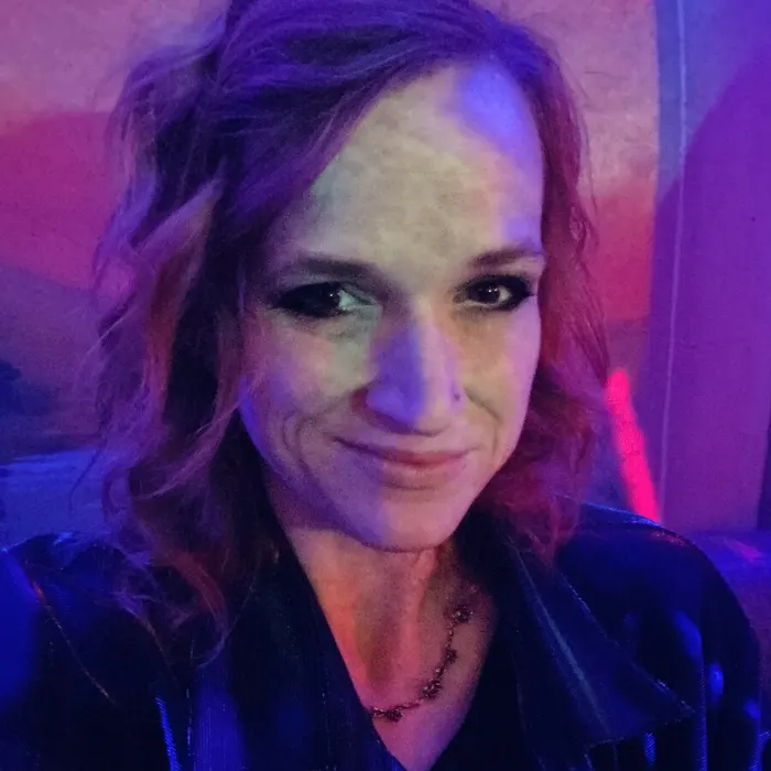 Meg at a heavy metal show, in a black jacket under purple and pink stage lights, with dark eye makeup and a half smile