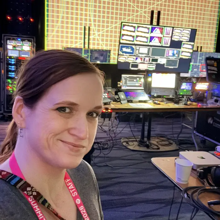 Meg smiling in a livestream control room, wearing a Summit staff lanyard, with production monitors behind her