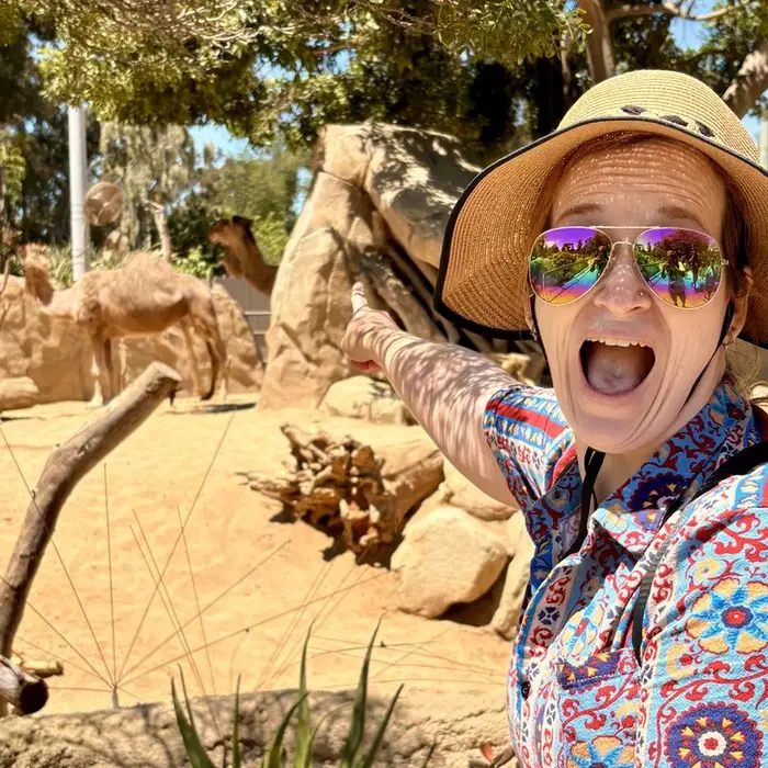 Meg in a sun hat and rainbow sunglasses, mouth open in delight, pointing at a camel behind her