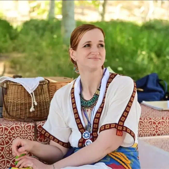 Meg in handmade Minoan-style garb, a white tunic with red woven trim and a beaded collar, sitting outdoors beside a wicker basket