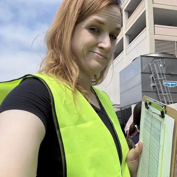 Meg in a yellow safety vest at a fire drill rally point, holding a clipboard with a checklist of names and giving the camera a sideways look