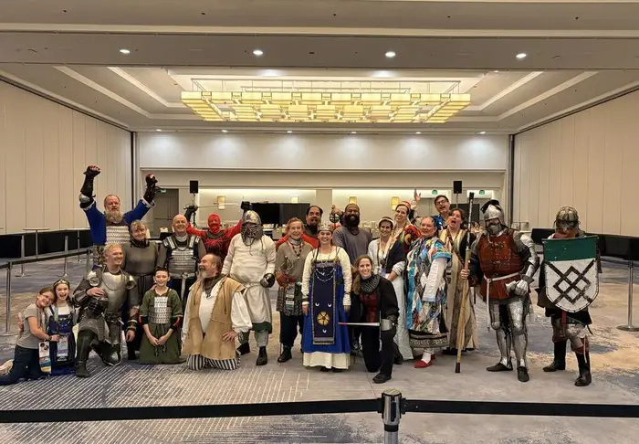 A large group of SCA fighters in armor and volunteers in period clothing posing in a convention hall after the combat demo