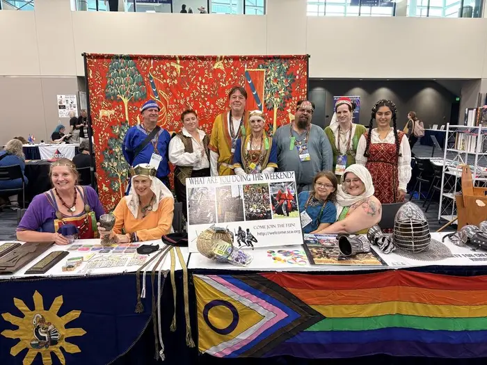 SCA volunteers in medieval clothing at a convention fan table, with a tapestry backdrop and a sign asking what you're doing this weekend